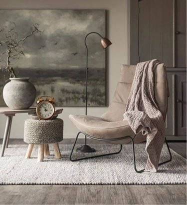 An empty chair with a sweater draped over it, with a stool, clock, vase, and landscape painting hung up in the background