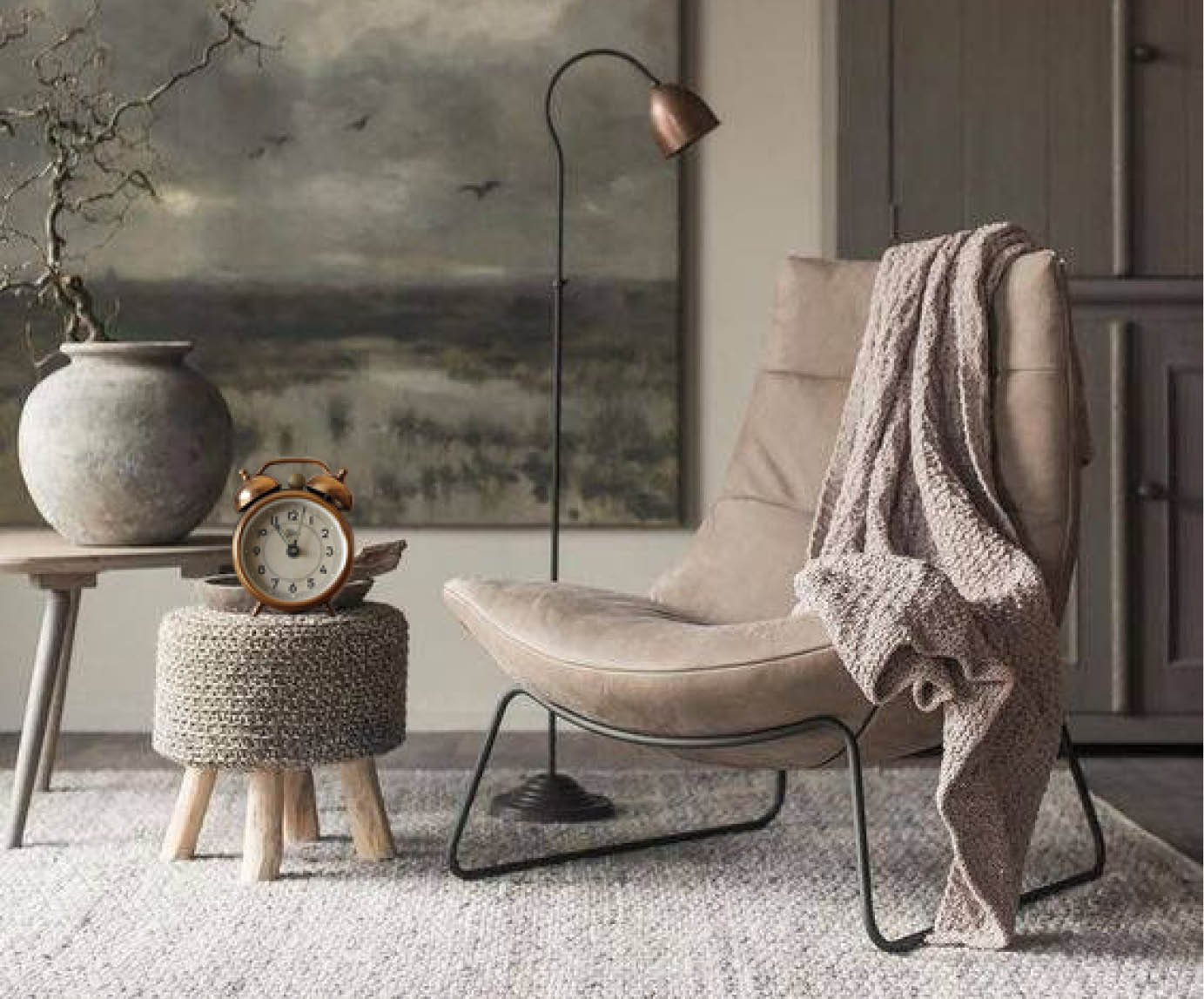 An empty chair with a sweater draped over it, with a stool, clock, vase, and landscape painting hung up in the background